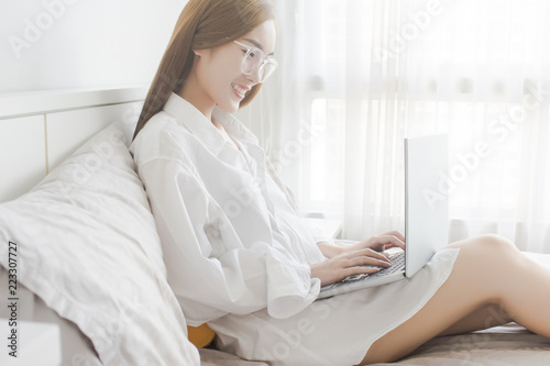  Happy casual beautiful Asian woman working on a laptop sitting on the bed in the house.