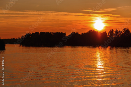Bright and colorful sunset on the river. The sun hides behind the trees in the evening. Glare of the setting sun on the water.