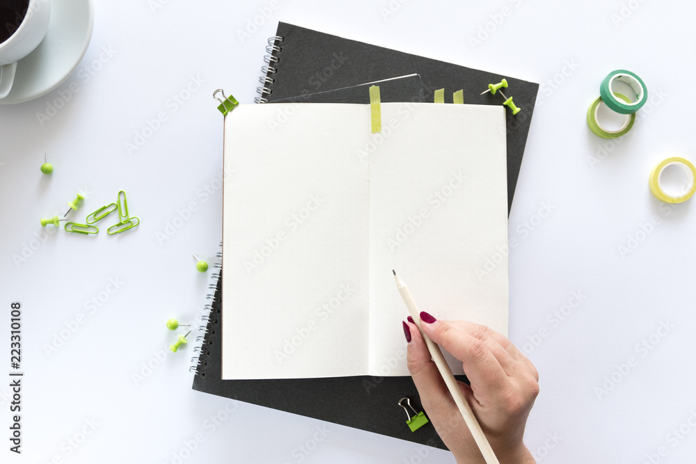 Top view over the working desk with notebook, cup fo coffee, paper clips and woman hand writing something. Copy space for text. Flat lay of a workplace on white background Top view over the working desk with notebook, cup fo coffee, paper clips and woman hand writing something. Copy space for text. Flat lay of a workplace on white background