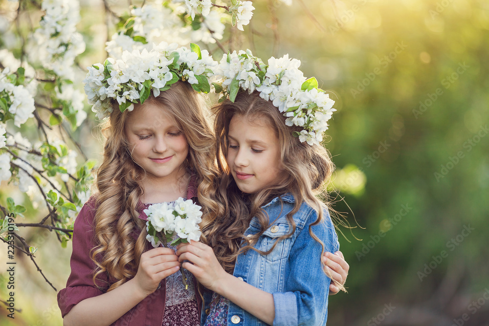 Obraz premium Two cute little girls friends with wreath of flowers in a blooming apple orchard