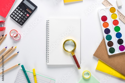 Top view over a school supplies as calculator, rulers, tapes, paper clips, notebooks and other stuff placed in a circle on a white background with notebook and magnificent glass in the middle of it. B