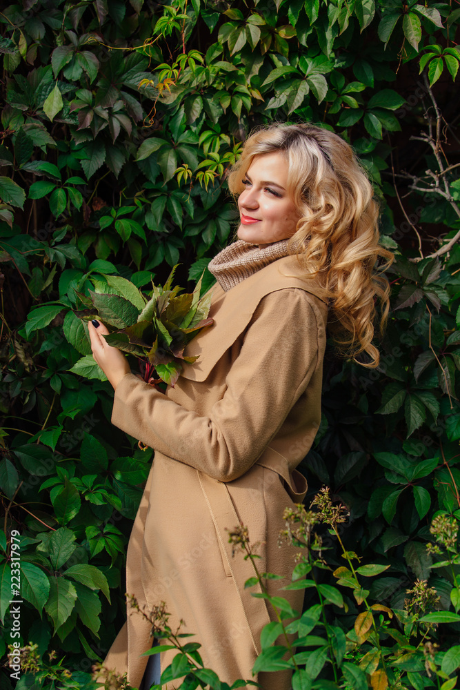 Young beautiful woman in sweater and coat stands next to the background of wild grapes holding bouquet of autumn leaves Young beautiful woman in sweater and coat stands next to the background of wild grapes holding bouquet of autumn leaves