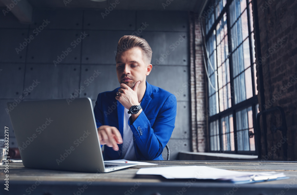 Young attractive business man in blue suit is sitting in the office at the desk with laptop and phone, marketing and communication management Young attractive business man in blue suit is sitting in the office at the desk with laptop and phone, marketing and communication management