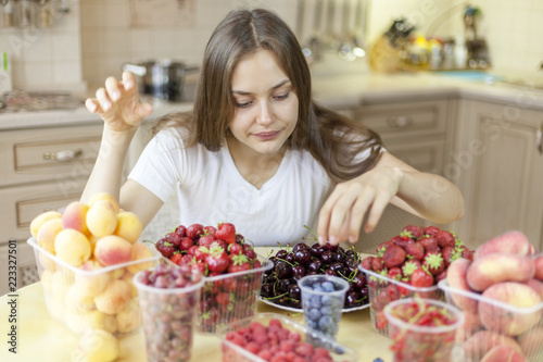 Beautiful young girl eats fruits and fresh berries in the kitchen