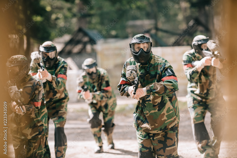 Fototapeta premium selective focus of paintball team in uniform and protective masks playing paintball with marker guns outdoors
