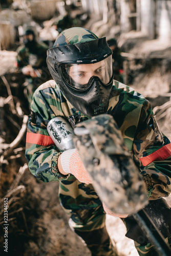 selective focus of male paintball player in goggle mask and camouflage aiming by paintball gun from ditch outdoors
