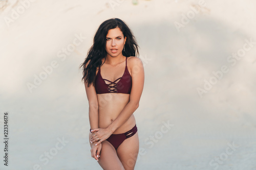 Seductive bright tanned girl in swimsuit posing on white sand, showing her beautiful slender figure and slim facial features, has a European appearance, curly dark, dark hair © romannoru