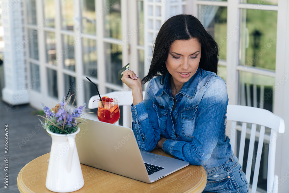 A beautiful tanned woman in a jeans shirt is sitting in a cafe, a laptop is open in front of her, a woman wants to buy something in an online store. A beautiful tanned woman in a jeans shirt is sitting in a cafe, a laptop is open in front of her, a woman wants to buy something in an online store.
