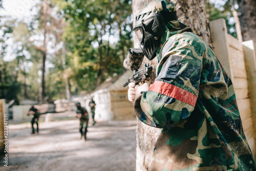 male paintballer in camouflage and protective mask holding marker gun and hiding behind tree while other team running behind outdoors photo