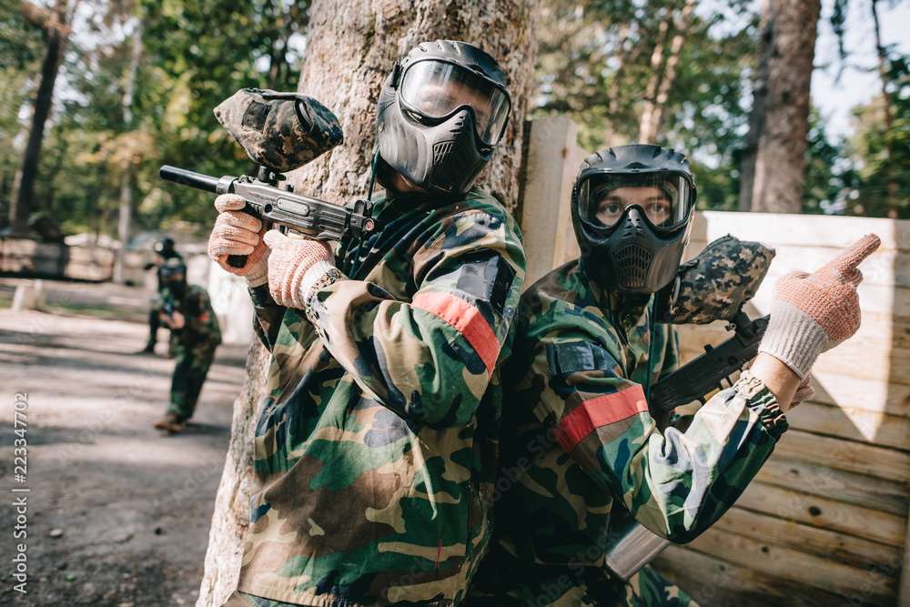 male paintball player in camouflage and goggle mask pointing by finger to his teammate with marker gun standing near tree outdoors