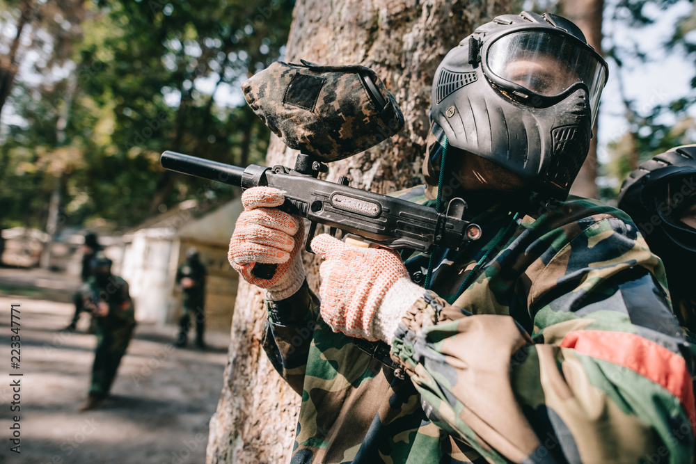 Fototapeta premium paintball players in camouflage and protective masks talking and hiding behind tree outdoors