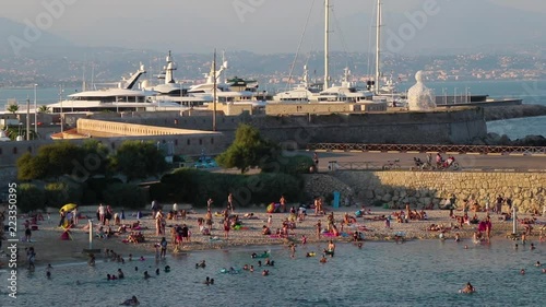 Crowded beach (Plage de la Gravette) near Port Vauban in Antibes, southern France. Boats in harbour, statue 