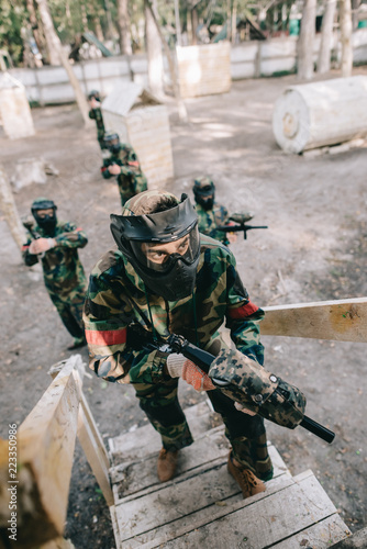 high angle view of male paintball player in goggle mask with marker gun on staircase while his team standing behind outdoors