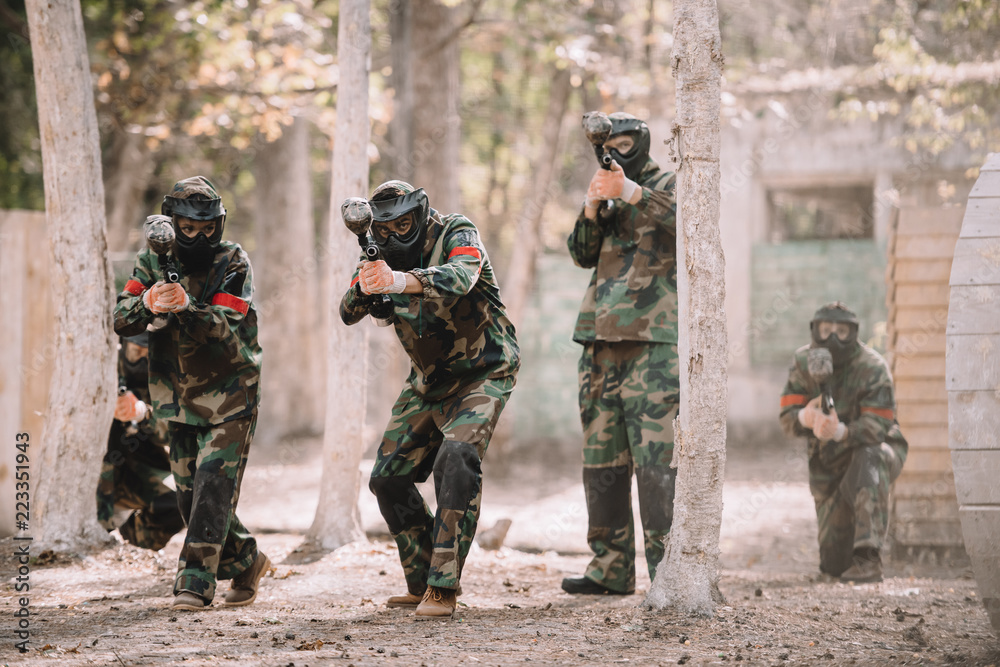 Fototapeta premium paintball team in uniform and protective masks running and aiming by paintball guns outdoors