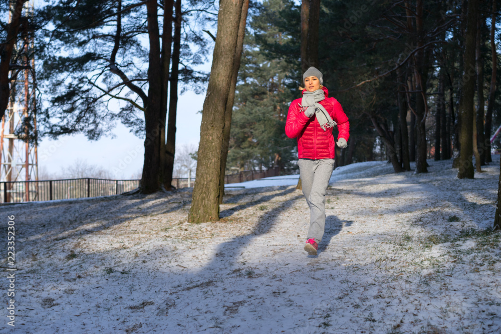 Female runner jogging in cold winter forest wearing warm sporty running clothing and gloves.