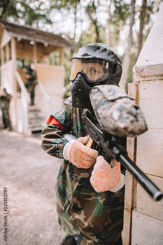 Fototapeta premium focused male paintballer in goggle mask covered by paintball splash aiming by marker gun outdoors