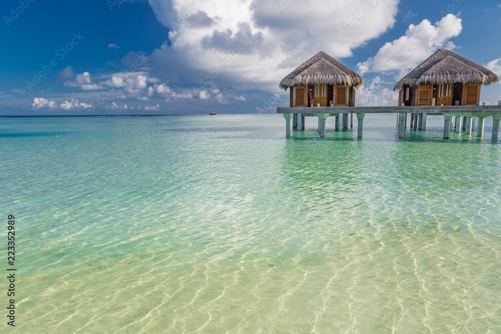 Obraz premium Water villas in lagoon, Maldives resort island in sunset. Detail of palm leaves on foreground. Vacation and beach relaxation, summer holidays background
