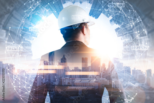 The double exposure image of the engineer standing back during sunrise overlay with cityscape image and futuristic hologram. The concept of engineering, construction, city life and future.