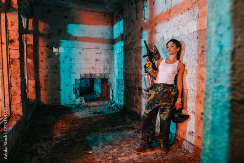 young female paintballer in camouflage and white singlet holding paintball gun and goggle mask in abandoned building