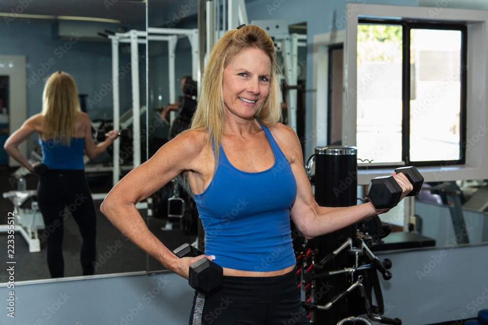 Fototapeta premium Strong and muscular middle age female showing impressive arms while posed with dumbbell weight in hand..