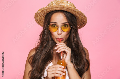 Photo closeup of charming woman 20s wearing sunglasses and straw hat drinking juice from glass bottle, isolated over pink background