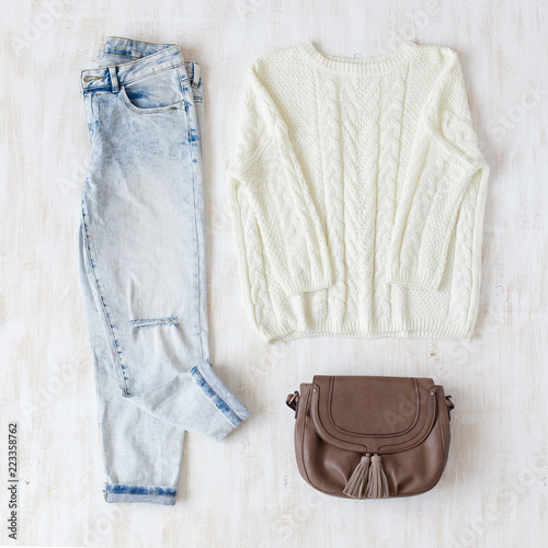 Light blue ripped boyfriend jeans, white oversize knitted sweater and small brown leather bag with tassels on white wooden background. Overhead view of woman's casual day outfits. Trendy hipster look.