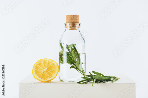 bottle of natural herbal essential oil with green twig and lemon on white cube