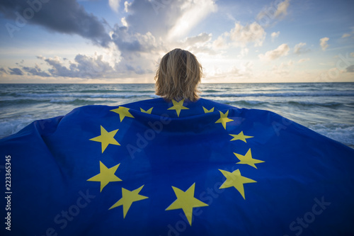 Man holding a fluttering iconic EU flag with circle of stars on beach with stormy turbulent seas in the channel at sunrise
