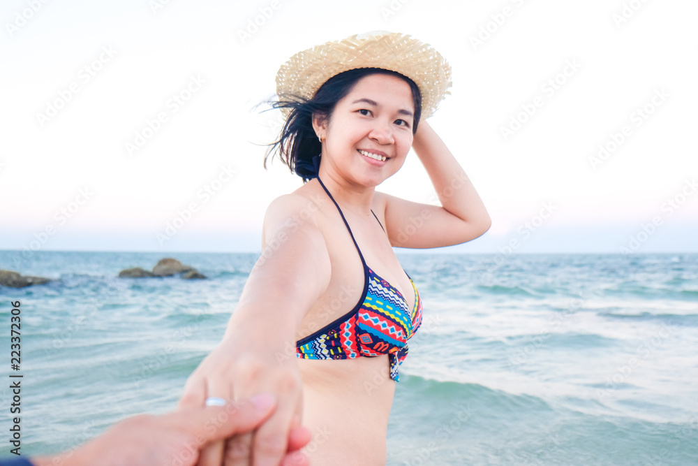 Asian girlfriend wear hat leading man hand to travel on sea beach
