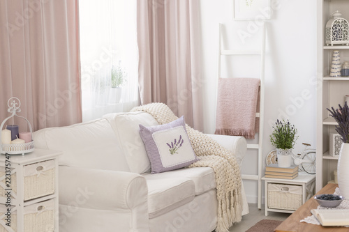 Pillows and blanket on white sofa in pink living room interior with drapes and flowers. Real photo