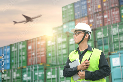Engineer foreman is standing with stacking of shipping containers in the depot with Air freight service at background for shipping, industry and logistics success concept.