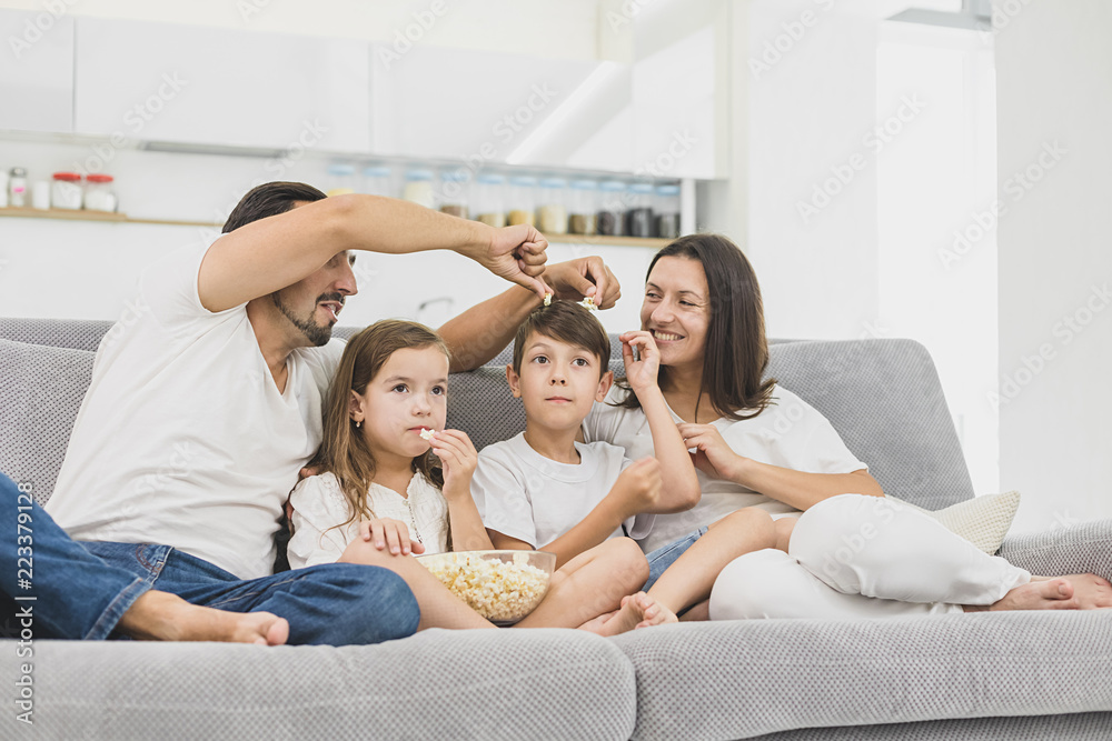 Beautiful young parents and their children are watching TV, eating popcorn and smiling while sitting on couch at home