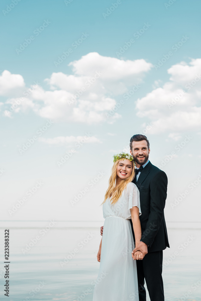 smiling wedding couple in suit and white dress holding hands on beach