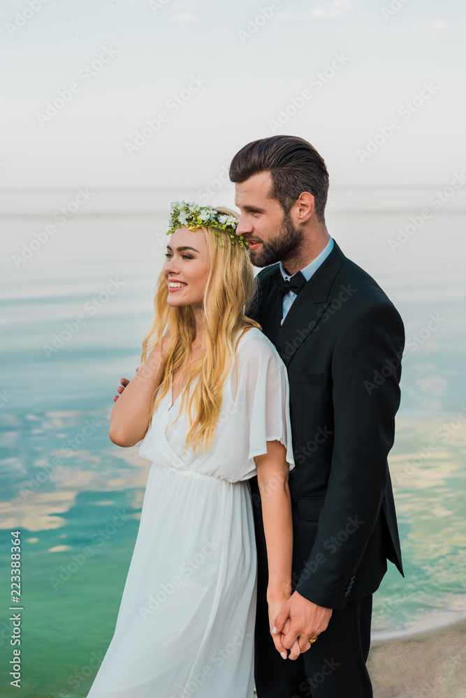 wedding couple in suit and white dress holding hands and looking away on beach wedding couple in suit and white dress holding hands and looking away on beach