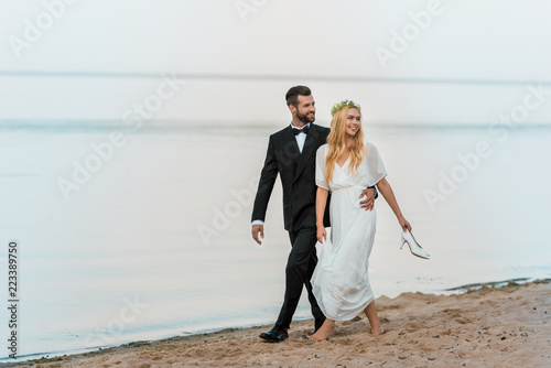 wedding couple hugging  walking on beach and looking away  bride holding high heels in hand