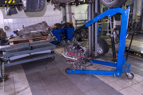 A sports car raised on a lift for repair and under it a detached engine suspended on a blue crane and a gear box on a lifting table in a vehicle repair shop