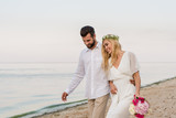 handsome groom hugging beautiful bride with wedding bouquet and they walking on beach