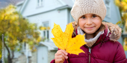 childhood, season and real estate concept - close up of happy little girl with fallen maple leaf in autumn over living house background outdoors