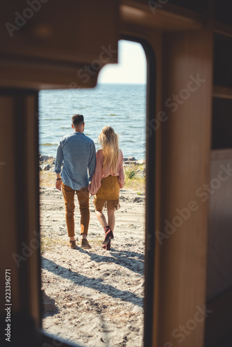 back view of hippie couple holding hands and walking near sea, from trailer door
