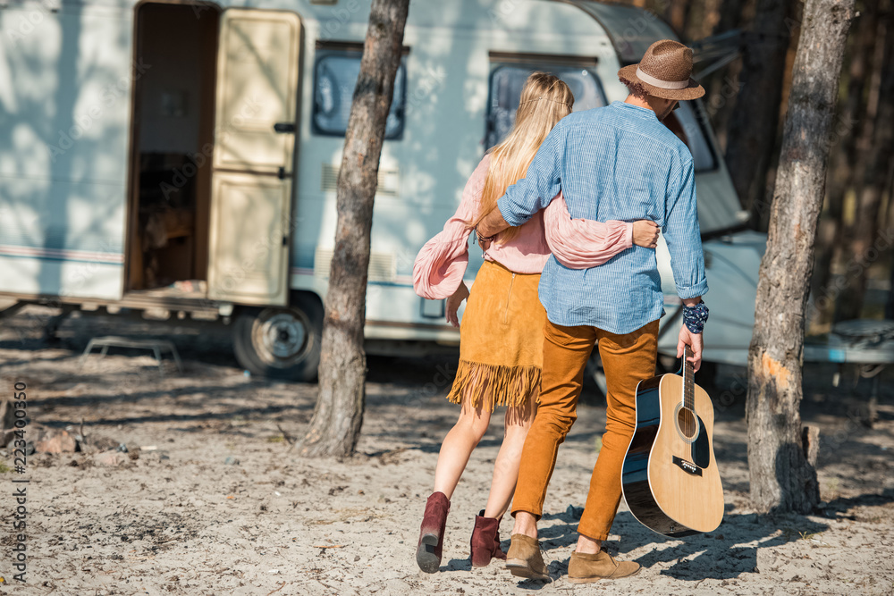 rear view of hippie couple hugging and walking with guitar near trailer