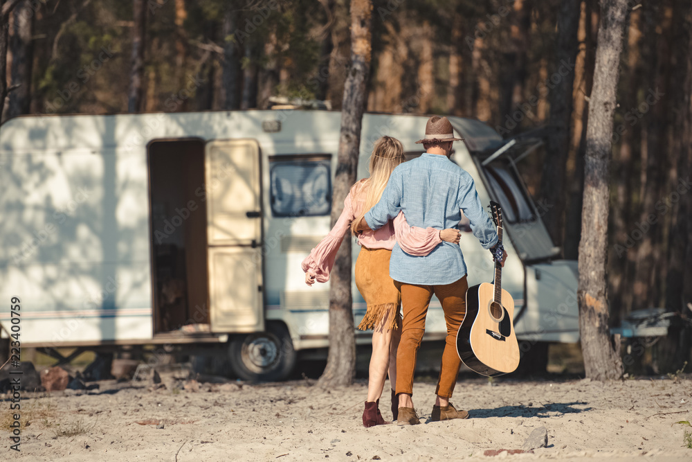 back view of hippie couple hugging and walking with acoustic guitar near campervan