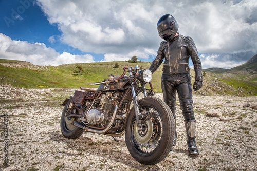 Biker with black leather clothes, standing near his custom rat motorcycle in a desolated mountain land. Post apocalyptic concept