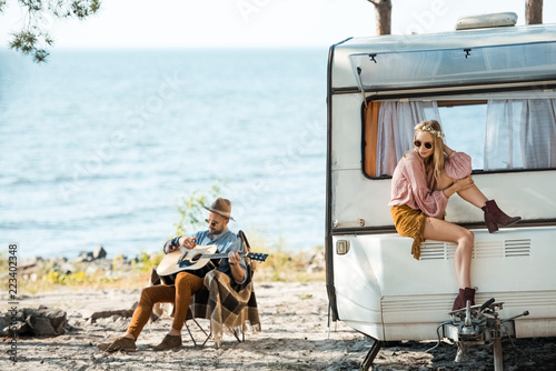 beautiful hippie girl sitting on campervan while man playing guitar near sea
