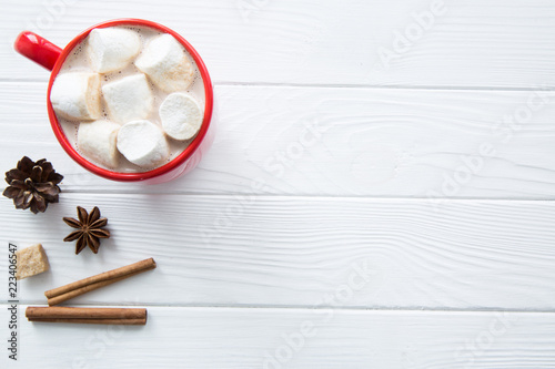 Christmas red cup with hot chocolate and marshmallow. Hot cocoa with milk, anise and cinnamon, brown sugar on white wooden rustic background, top view. Copy space.