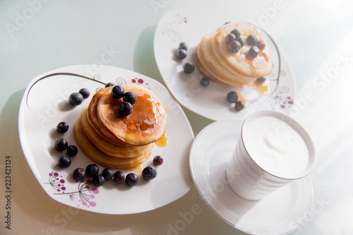 Homemade pancake plates topped by maple syrup, cinnamon and blueberries on white plates with cup of delicious coffee topped with cream, yummy and sweet breakfast table for two