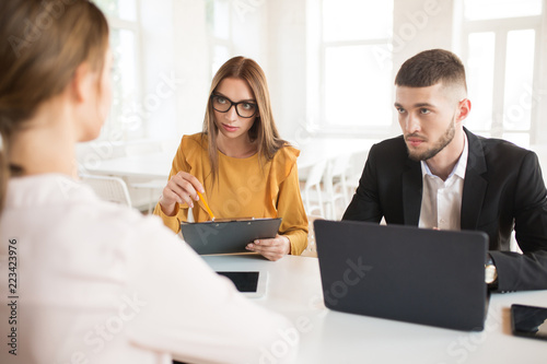 Young business man with laptop and business woman in eyeglasses thoughtfully looking at applicant. Young employers spending job interview in office
