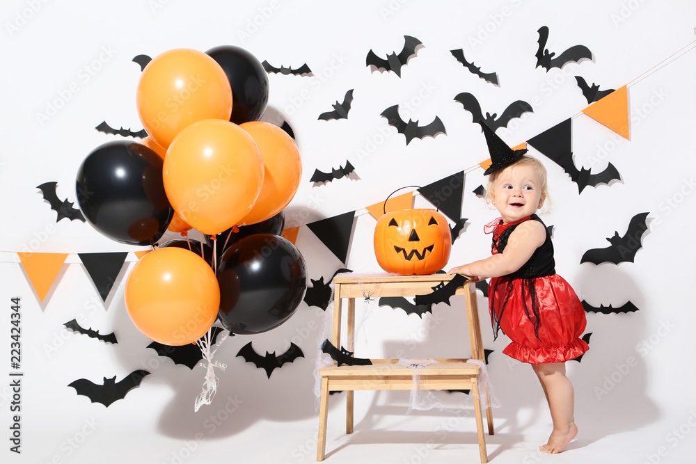 Fototapeta premium Baby girl in halloween costume with balloons and pumpkin bucket on white background