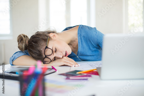 Young tired woman with eyeglasses on head sleeping on desk while working in modern office