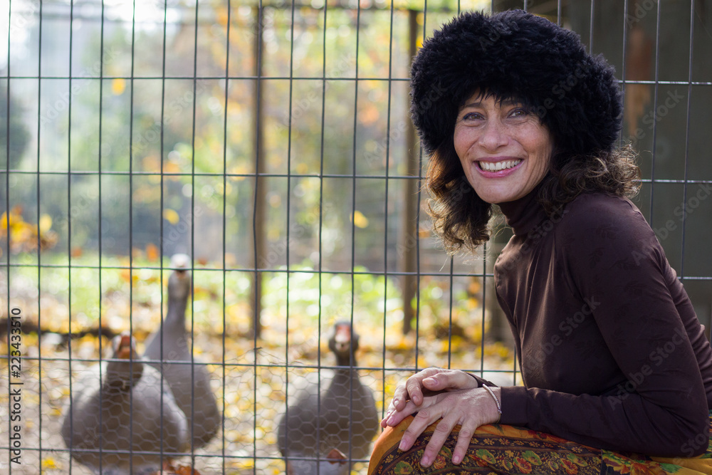Elegant middle aged woman posing happily next to geese on the other side of  fence at
