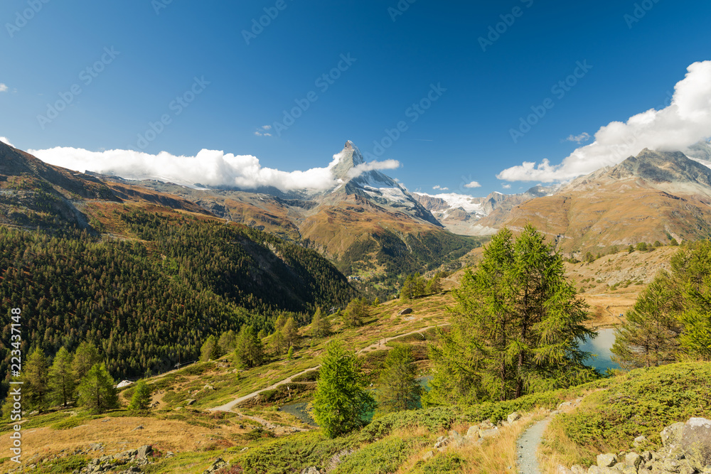 Scenery summer view on snowy Matterhorn peak in sunny day with pine trees forest, mountains and blue sky with some clouds in background, Switzerland